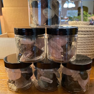 May include: A stack of clear plastic jars with black lids, each filled with various colorful gemstones. The gemstones include pink, white, and dark-colored stones. The jars are arranged on a wooden surface.