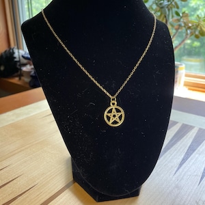 May include: A gold-colored necklace with a pentagram pendant. The pendant is a five-pointed star within a circle. The necklace is displayed on a black velvet bust against a background of a wooden table and a window.