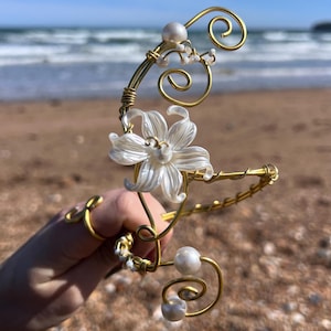 May include: A gold-toned wire headpiece featuring a white flower and faux pearls. The floral design is accented with delicate swirls and small pearl embellishments. The headpiece is held up against a beach backdrop.