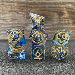 May include: A set of six polyhedral dice for tabletop games. The dice are clear with blue and gold accents, featuring gold numerals. The dice are arranged on a dark surface with a wooden background.