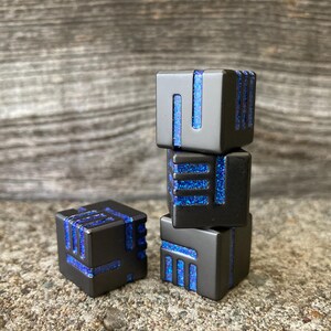 May include: Four black square dice with blue glitter accents. The dice are stacked in a tower of three, with one die separate. The dice have a modern, geometric design.