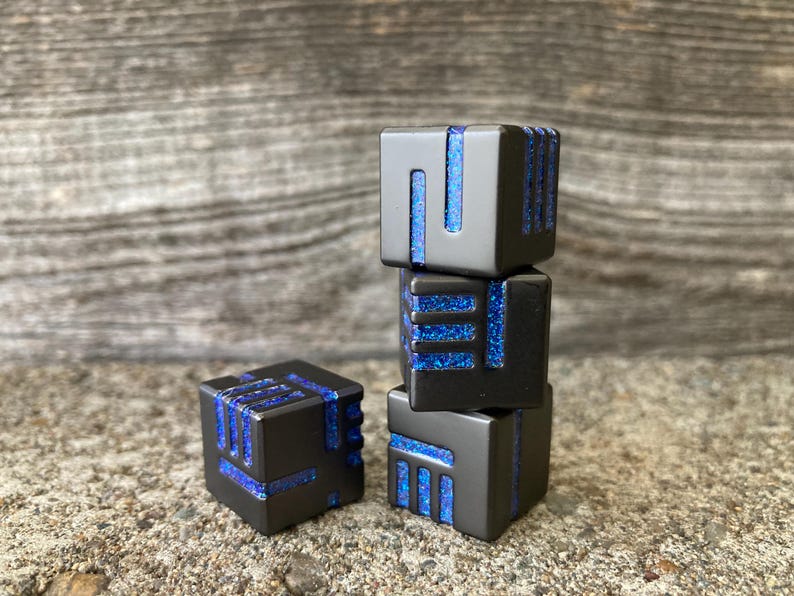 May include: Four black square dice with blue glitter accents. The dice are stacked in a tower of three, with one die separate. The dice have a modern, geometric design.