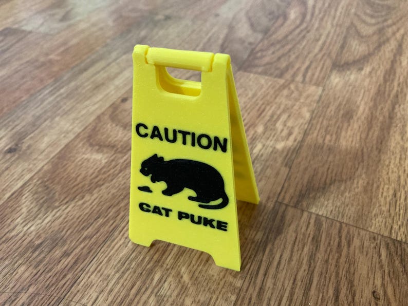 May include: A bright yellow, A-frame caution sign featuring a black silhouette of a cat and the black text CAUTION CAT PUKE. The sign is constructed with a hinged top handle and stands on four small feet, mimicking a standard floor warning sign.