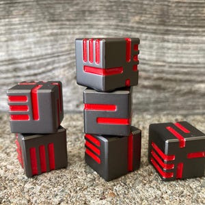 May include: Six dark gray, cube-shaped objects with red accents are stacked and arranged on a textured surface. The cubes have a geometric design with red lines and shapes. The overall aesthetic is modern and minimalist.