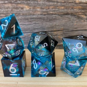 May include: A collection of six polyhedral dice in various shapes, including a d4, d6, d8, d10, d12, and d20. The dice are a translucent teal color with silver numbering and glitter inclusions. The dice are arranged on a wooden surface.
