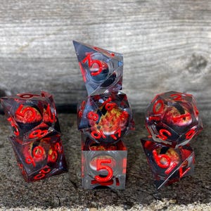 May include: A collection of clear, multi-sided dice with red numbers and a fiery interior design. The dice are arranged on a textured surface, showcasing their transparent quality and intricate details. The dice are designed for tabletop games.