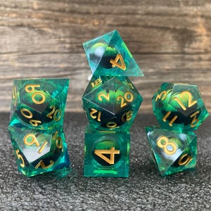 May include: A set of seven polyhedral dice in various shapes, including a d4, d6, d8, d10, d12, d20, and a percentile die. The dice are transparent teal with gold numbering and a swirling green and black interior. The dice are arranged on a dark gray surface.