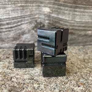 May include: Three dark gray, cube-shaped objects with a geometric design. One cube is separate, while the other two are stacked. The cubes have a metallic appearance and are set against a textured, neutral background.
