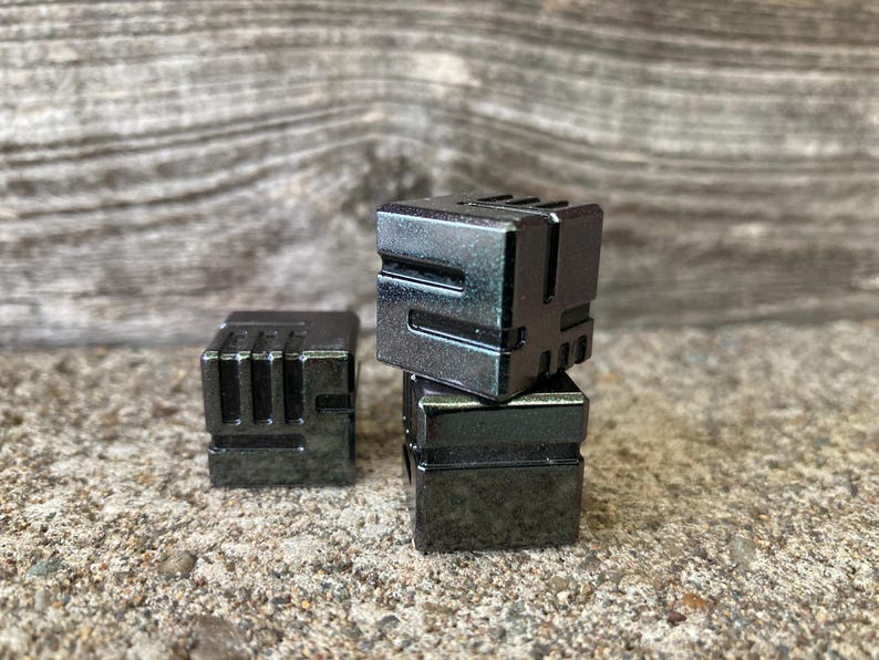May include: Three dark gray, cube-shaped objects with a geometric design. One cube is separate, while the other two are stacked. The cubes have a metallic appearance and are set against a textured, neutral background.
