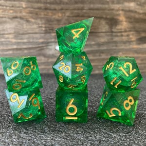 May include: Set of seven green and gold polyhedral dice. The dice are transparent with gold numbering. The dice are arranged in a stack on a dark gray surface. The dice are for tabletop games.