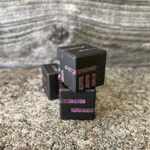 May include: Three dark gray, cube-shaped objects with glitter accents. The glitter is a mix of pink and gold. The cubes are stacked on top of each other, with one slightly offset. The background is a textured, gray surface.