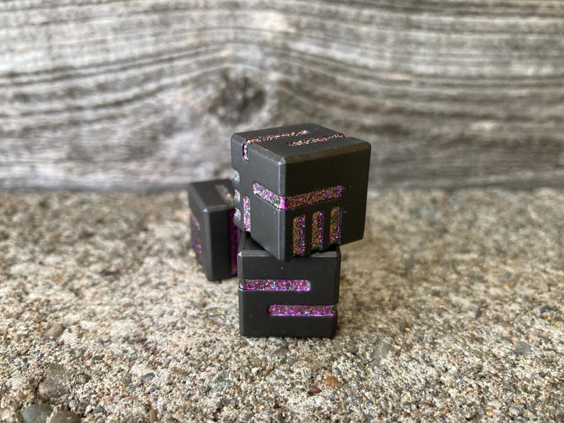 May include: Three dark gray, cube-shaped objects with glitter accents. The glitter is a mix of pink and gold. The cubes are stacked on top of each other, with one slightly offset. The background is a textured, gray surface.