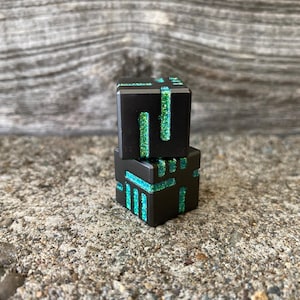 May include: Two black, geometric dice with glitter accents in teal and green. The dice are stacked on top of each other, with the top die slightly offset. The background is a textured, gray surface.