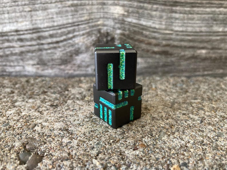 May include: Two black, geometric dice with glitter accents in teal and green. The dice are stacked on top of each other, with the top die slightly offset. The background is a textured, gray surface.