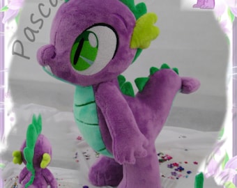 Può includere: Un giocattolo di peluche di drago viola e verde con una grande cresta verde e una lunga coda. Il drago è in piedi su una superficie bianca con coriandoli colorati.