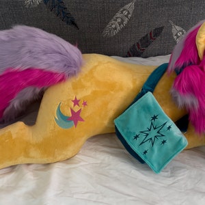 Könnte beinhalten: Ein Plüschpferd in liegender Position, mit gelbem Körper, rosa und lila Mähne und Schweif sowie einer türkisfarbenen Umhängetasche. Das Pony hat ein Mond- und Stern-Design an der Flanke. Die Umhängetasche hat ein Sternenmuster.