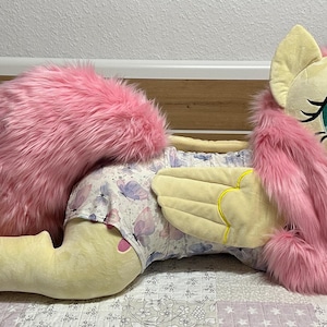 Puede incluir: Un peluche grande de poni con piel amarilla pálida, pelo largo de piel sintética rosa y cola rosa. El poni tiene ojos grandes de color verde azulado y alas pequeñas con costuras amarillas. Lleva un pañal blanco con estampado floral.