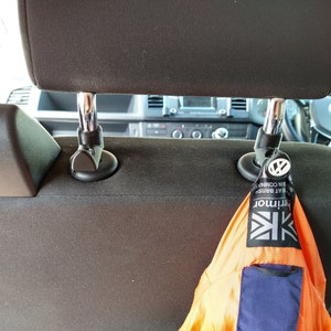 May include: Two black car headrest hooks with a bright orange and blue jacket hanging from one of the hooks. The jacket has a white and black logo that says "Kithmor" and "Great British Countrywear" with a Union Jack flag.