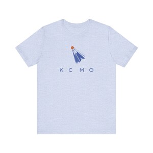Può includere: Una t-shirt blu chiaro con una grafica di volano da badminton blu e arancione e il testo "KCMO" stampato sul davanti.