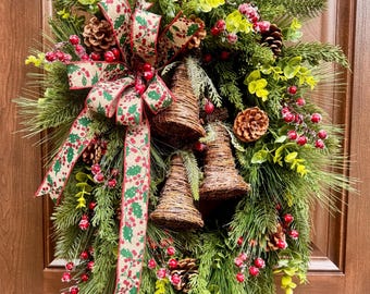 May include: A holiday wreath featuring green pine needles, brown pinecones, red berries, and light green foliage. Three woven brown bells hang in the center, accented by a tan ribbon with a red and green holly pattern. The text Caleb's Mom's Shop is visible.