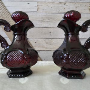 May include: Two red glass pitchers with a textured design and a handle. The pitchers have a round body and a small spout.