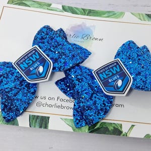 May include: Two hair bows made of bright blue glitter fabric, each featuring a shield-shaped emblem with the text State of Origin NSW Blues and a rugby ball graphic. The bows are layered with a textured, sparkling finish and a central knot detail.