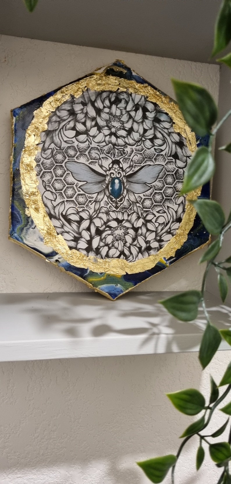May include: A hexagonal resin art piece with a gold leaf border. The center features a black and white illustration of a bee with a blue gemstone on its body, surrounded by flowers and a honeycomb pattern.