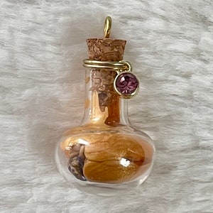 May include: A small glass bottle with a cork stopper and a gold wire around the neck. The bottle is filled with a golden liquid and a small purple gemstone charm hangs from the wire.