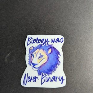 Peut inclure: Un autocollant découpé présentant une illustration stylisée d'un lion avec une crinière bleue et violette et un visage crème. Le texte "Biology was Never Binary" est écrit en écriture cursive bleu foncé au-dessus et sous la tête du lion.