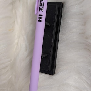Puede incluir: Un bol&iacute;grafo retr&aacute;ctil de color morado claro con un clip de bolsillo y el texto negro HI ZEV! impreso verticalmente a lo largo del cuerpo. El bol&iacute;grafo est&aacute; colocado contra un soporte rectangular negro con acabado texturizado.