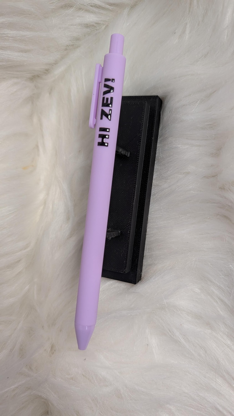 Puede incluir: Un bol&iacute;grafo retr&aacute;ctil de color morado claro con un clip de bolsillo y el texto negro HI ZEV! impreso verticalmente a lo largo del cuerpo. El bol&iacute;grafo est&aacute; colocado contra un soporte rectangular negro con acabado texturizado.