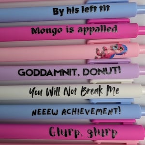 Puede incluir: Una colecci&oacute;n de diez bol&iacute;grafos retr&aacute;ctiles en varios colores pastel, incluyendo rosa, azul, morado y blanco. Cada bol&iacute;grafo presenta texto impreso en negro o un peque&ntilde;o gr&aacute;fico. Las frases incluyen IWKYM, By his left tit, Mongo is appalled, GODDAMNIT, DONUT!, You Will Not Break Me, NEEEW ACHIEVEMENT!, Glurp, glurp, Mama Needs a Night Off y HI, ZEV!.