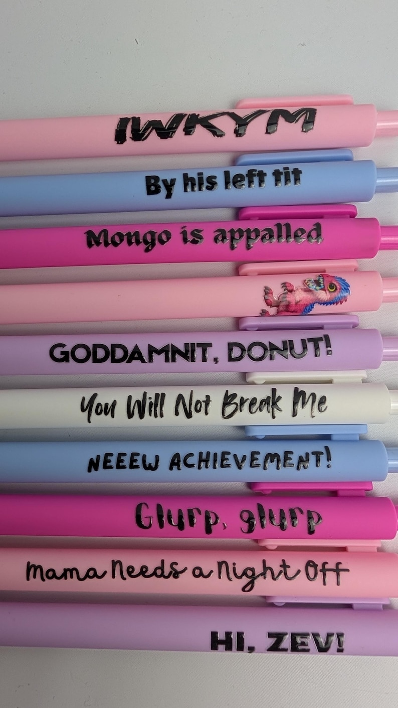 Puede incluir: Una colecci&oacute;n de diez bol&iacute;grafos retr&aacute;ctiles en varios colores pastel, incluyendo rosa, azul, morado y blanco. Cada bol&iacute;grafo presenta texto impreso en negro o un peque&ntilde;o gr&aacute;fico. Las frases incluyen IWKYM, By his left tit, Mongo is appalled, GODDAMNIT, DONUT!, You Will Not Break Me, NEEEW ACHIEVEMENT!, Glurp, glurp, Mama Needs a Night Off y HI, ZEV!.