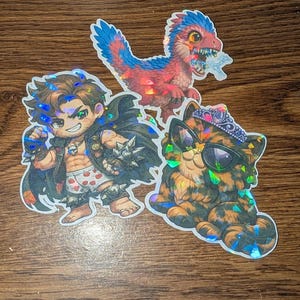 Puede incluir: Tres pegatinas holográficas sobre una superficie de madera. Una pegatina presenta un hombre de dibujos animados con una capa y una armadura puntiaguda. Otra es un dinosaurio rojo y azul. La tercera es un gato con gafas de sol y una tiara.