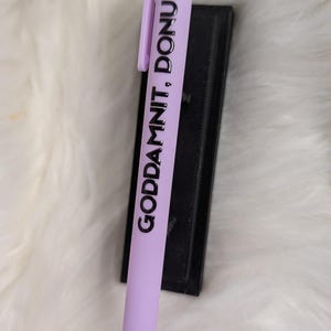 Puede incluir: Un bol&iacute;grafo retr&aacute;ctil de color morado claro con un clip y el texto impreso en negro "GODDAMNIT, DONUT!" a lo largo del cuerpo. El bol&iacute;grafo tiene un acabado mate suave y un dise&ntilde;o sencillo y funcional.
