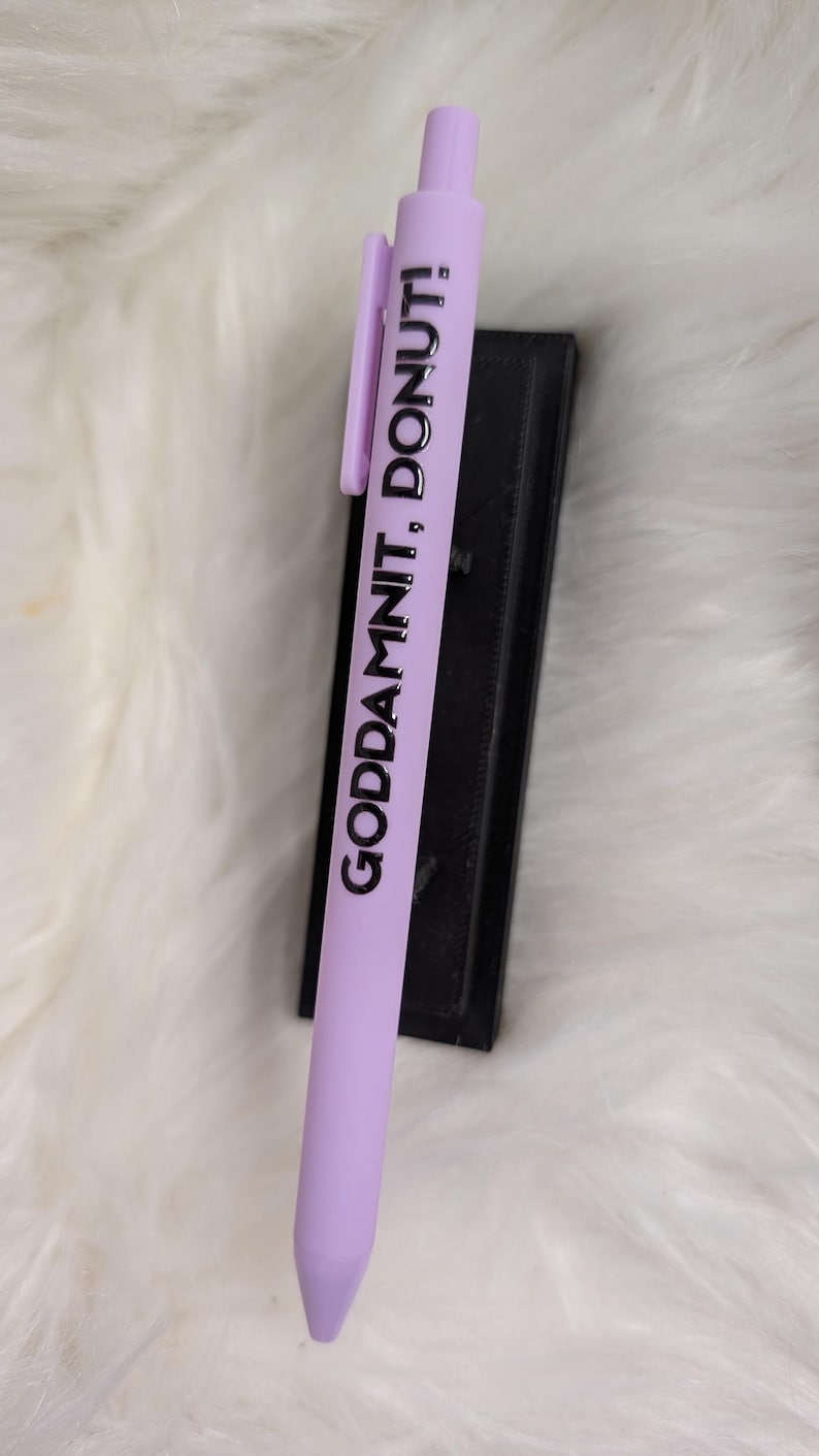 Puede incluir: Un bol&iacute;grafo retr&aacute;ctil de color morado claro con un clip y el texto impreso en negro "GODDAMNIT, DONUT!" a lo largo del cuerpo. El bol&iacute;grafo tiene un acabado mate suave y un dise&ntilde;o sencillo y funcional.