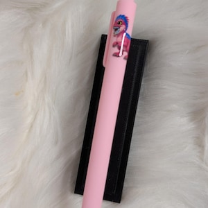 Puede incluir: Un bol&iacute;grafo retr&aacute;ctil de color rosa claro con un peque&ntilde;o gr&aacute;fico de un dinosaurio rosa con pelo azul en el cuerpo. El bol&iacute;grafo tiene un clip rosa a juego y un mecanismo de clic, presentado sobre una base rectangular negra.
