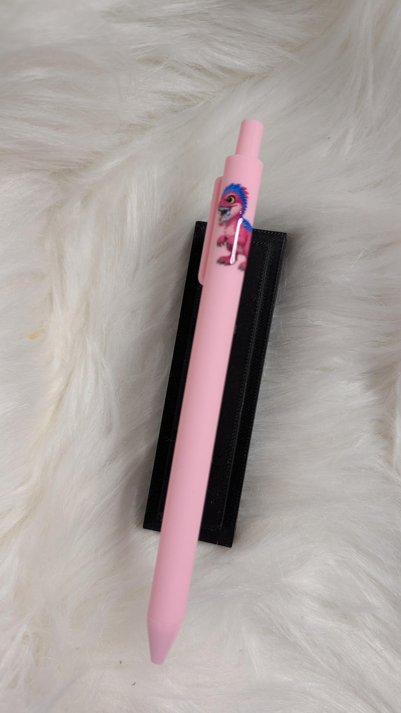 Puede incluir: Un bol&iacute;grafo retr&aacute;ctil de color rosa claro con un peque&ntilde;o gr&aacute;fico de un dinosaurio rosa con pelo azul en el cuerpo. El bol&iacute;grafo tiene un clip rosa a juego y un mecanismo de clic, presentado sobre una base rectangular negra.