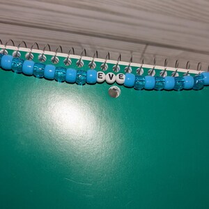 May include: A beaded keychain featuring translucent light blue plastic pony beads strung on a white cord. The center of the strand includes three white cube beads with the black printed letters E, V, and E, and a metal lobster clasp at one end.