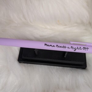 Puede incluir: Un bol&iacute;grafo retr&aacute;ctil de color morado claro con el texto "Mama Needs a Night Off" impreso en negro en el cuerpo, colocado horizontalmente sobre un peque&ntilde;o soporte de exhibici&oacute;n rectangular de pl&aacute;stico negro.