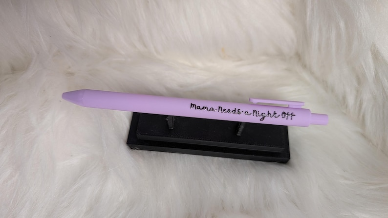 Puede incluir: Un bol&iacute;grafo retr&aacute;ctil de color morado claro con el texto "Mama Needs a Night Off" impreso en negro en el cuerpo, colocado horizontalmente sobre un peque&ntilde;o soporte de exhibici&oacute;n rectangular de pl&aacute;stico negro.