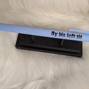 Puede incluir: Un bol&iacute;grafo retr&aacute;ctil de color azul claro con el texto negro By his left tit impreso en el cuerpo. El bol&iacute;grafo se muestra horizontalmente sobre un peque&ntilde;o soporte rectangular negro con dos soportes verticales que lo sostienen.
