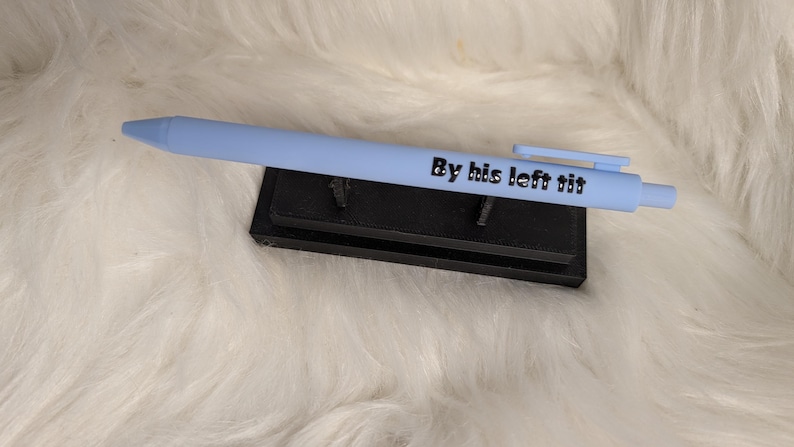 Puede incluir: Un bol&iacute;grafo retr&aacute;ctil de color azul claro con el texto negro By his left tit impreso en el cuerpo. El bol&iacute;grafo se muestra horizontalmente sobre un peque&ntilde;o soporte rectangular negro con dos soportes verticales que lo sostienen.