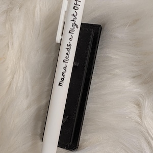 Puede incluir: Un bol&iacute;grafo retr&aacute;ctil blanco con un clip blanco y el texto impreso en negro "Mama Needs a Night Off" en una fuente cursiva, apoyado contra un soporte rectangular negro con una superficie texturizada.