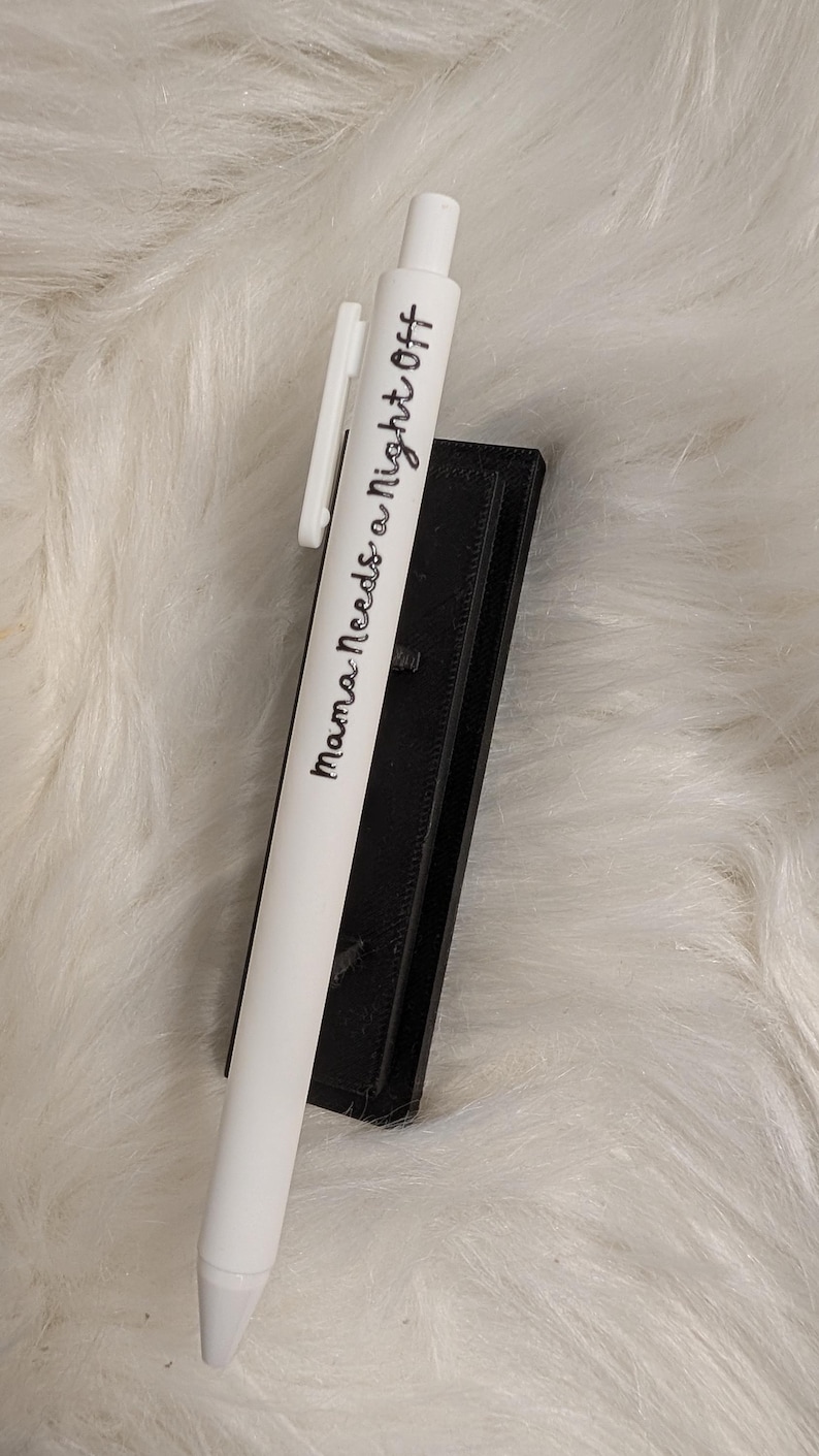 Puede incluir: Un bol&iacute;grafo retr&aacute;ctil blanco con un clip blanco y el texto impreso en negro "Mama Needs a Night Off" en una fuente cursiva, apoyado contra un soporte rectangular negro con una superficie texturizada.