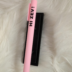 Puede incluir: Un bol&iacute;grafo retr&aacute;ctil de color rosa claro con el texto en negro HI ZEV! impreso verticalmente en el cuerpo. El bol&iacute;grafo incluye un clip de bolsillo rosa a juego y una punta c&oacute;nica, apoyado sobre un soporte rectangular negro.