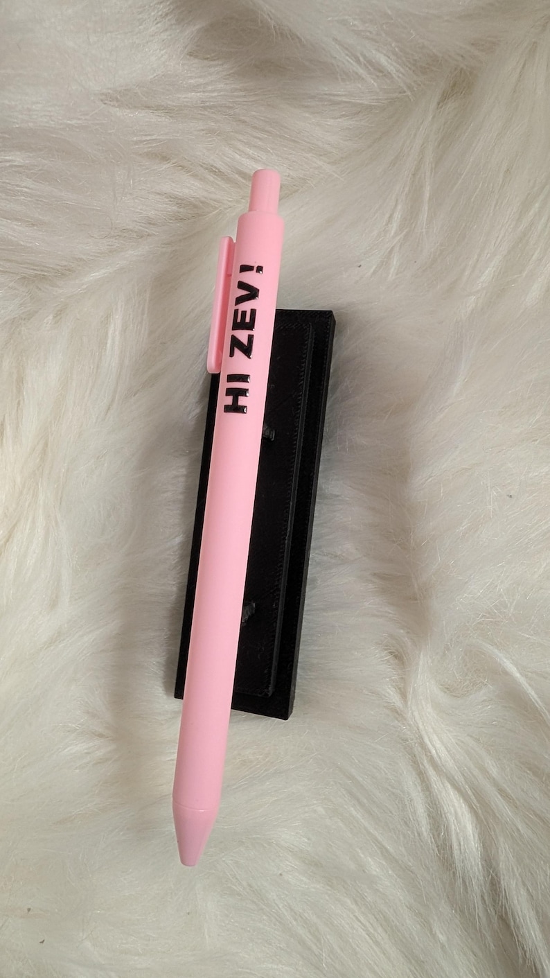 Puede incluir: Un bol&iacute;grafo retr&aacute;ctil de color rosa claro con el texto en negro HI ZEV! impreso verticalmente en el cuerpo. El bol&iacute;grafo incluye un clip de bolsillo rosa a juego y una punta c&oacute;nica, apoyado sobre un soporte rectangular negro.