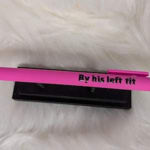 Puede incluir: Un bol&iacute;grafo retr&aacute;ctil de color rosa brillante con un clip de bolsillo y el texto negro "By his left tit" impreso claramente en el cuerpo. El bol&iacute;grafo descansa horizontalmente sobre un peque&ntilde;o soporte de exhibici&oacute;n negro.