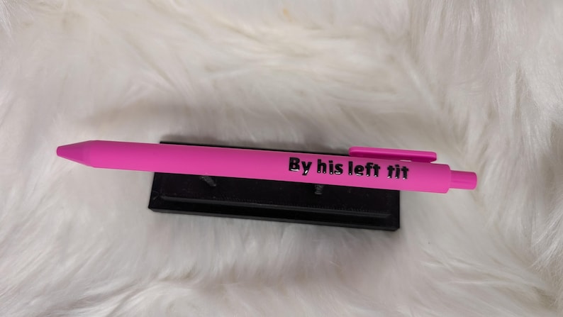 Puede incluir: Un bol&iacute;grafo retr&aacute;ctil de color rosa brillante con un clip de bolsillo y el texto negro "By his left tit" impreso claramente en el cuerpo. El bol&iacute;grafo descansa horizontalmente sobre un peque&ntilde;o soporte de exhibici&oacute;n negro.