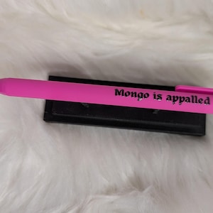 Puede incluir: Un bol&iacute;grafo retr&aacute;ctil de color rosa brillante con el texto en negro Mongo is appalled impreso en el cuerpo. El bol&iacute;grafo incluye un clip de bolsillo rosa a juego y est&aacute; colocado horizontalmente sobre un soporte negro.
